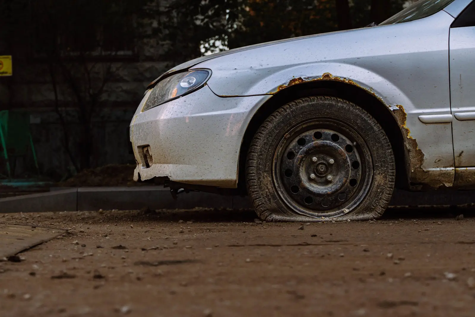 Flat tire on an older silver sedan parked on dirt